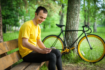 A young man sitting on a bench in a public Park communicates via mobile communication. In the background with his bike. Among trees and vegetation.