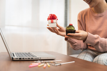 Asian woman celebrating birthday through video call online holding gift box and cupcake on hand while isolated stay at home