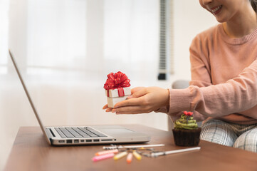 Asian woman celebrating birthday through video call online holding gift box and cupcake on hand while isolated stay at home