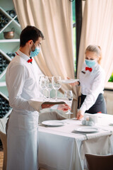 A waiter in a medical protective mask serves a table in the restaurant. Employees of a restaurant or hotel in protective masks. The End Of Quarantine.