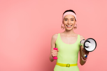 happy sportswoman holding dumbbell and loudspeaker on pink