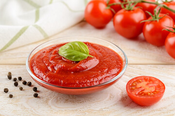 Tasty thick tomato ketchup in a glass bowl against ripe red cherry tomatoes on a twig over white wood table. Spices, seasonings and sauces.