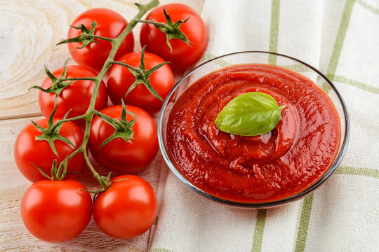 Tasty thick tomato ketchup in a glass bowl and ripe red cherry tomatoes on a twig over white wood kitchen table. Spices, seasonings and sauces.