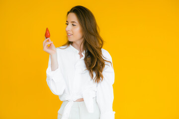 Fototapeta na wymiar Freshness comes in red. Close-up of beautiful woman holding strawberry in her hand and tasting it while standing against yellow background
