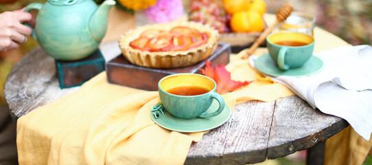 Autumn picnic. Tea party with beautiful kettle, cups at wooden table in garden. Harvest festival. Honey with stick, spoon, apple pie, persimmons, grapes, maple leaf, flowers, yellow linen tablecloth