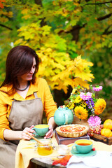 Autumn picnic. Woman in yellow dress and linen apron drinks tea from cup at wooden table in garden. Beautiful kettle, tablecloth, honey with spoon, apple pie, harvest, persimmon, grapes, maple leaf