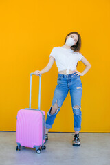 Beautiful girl in a white summer dress with a suitcase. Medical mask on the face. Closed borders during coronavirus. Tourist on a yellow background. Ban on travel to another country due to the virus