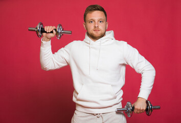 Fototapeta na wymiar A bearded man with a red beard and mustache, a blond man, dressed in sportswear, with joy on his face, lifts a dumbbell with one hand without effort, on a red background. Feels the power