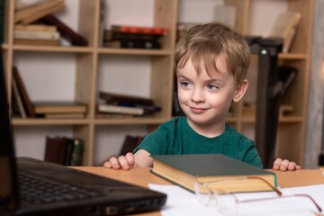 a three year old boy sits at a table and watches online lessons. a child on distance learning during quarantine. bookcase on the background. The opened book lies nearby.