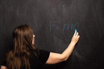 The physical formula of Einstein's theory is written by a young girl in blue chalk on a blackboard. FMC2 is written by a science teacher or student in the classroom.
