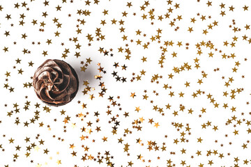 Delicious chocolate cupcake with chocolate icing and sprinkled gold sparkles on white background with golden star shaped confetti. Holiday concept. Flat lay style. Place for text.