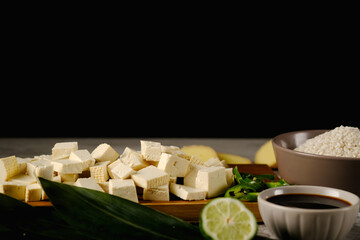 Fresh tofu pieces on bamboo leaves, on a wooden board near soy sauce, rice, ginger and lime.On a black background.Space for text.