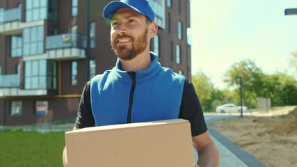 Close up handsome young happy smiling delivery man holding cardboard walking box at street outdoor shopping package portrait postman slow motion