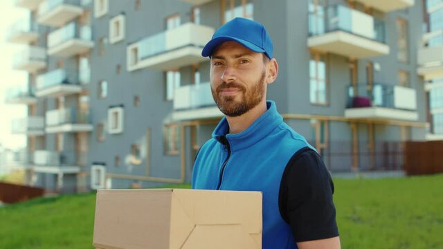 Handsome young happy smiling delivery man holding cardboard box stand look at camera at street outdoor shopping package postman slow motion