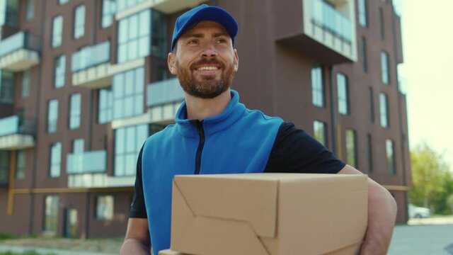 At sunlight attractive young happy smiling delivery man holding cardboard box stand look at camera at street outdoor shopping package postman slow motion