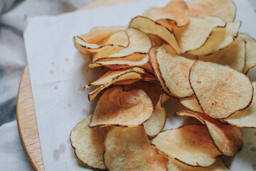 Fresh homemade deep fried crispy potato chips in white box on a wooden tray, top view. Salty crisps scattered on a table for a tasty snack break and party movie time.