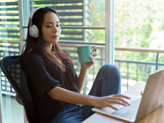 Female relaxed woman with headphone using laptop and holding a coffee cup in bed room