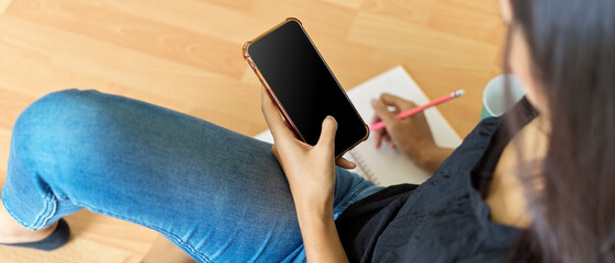A woman take note on blank notebook while looking information on smartphone