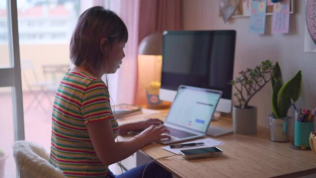young asian malaysian chinese woman working from home answering a call and typing on the laptop sitting on a desk from the side