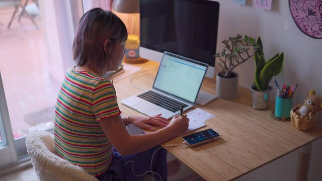 young asian malaysian chinese woman working from home answering a call and taking notes sitting on a desk from the side