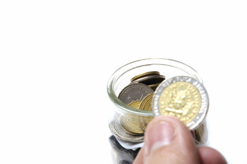 Glass jar with many Coins, hand holding a coin against white background