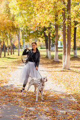 Fototapeta premium Ballerina with Dalmatian dog in the Park. Woman ballerina in a white ballet skirt and black leather jacket dancing in pointe shoes in autumn park with her spotty dalmatian dog.
