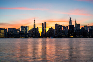 Manhattan Midtown skyline view. Dark silhouettes of Empire State building, Chrysler building, American Copper Buildings, Headquarters of the United Nations. Sunset, orange sky, reflections in water