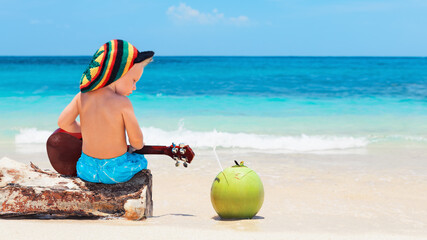 Little baby in rasta hat play reggae music on Hawaiian ukulele, enjoy relaxing on ocean beach. Kids...