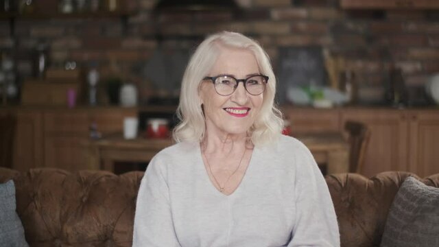 Elderly, beautiful woman with gray hair smiling looking at the camera. Portrait of a 70 year old grandmother with glasses. Happy senior citizen. An elderly, business woman sits on a sofa while being