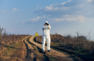 Obraz premium Scientist in radiation suit and gas mask showing that entrance on territory is forbidden, standing near yellow triangle with skull and crossbones sign and warning about poisonous substances and danger