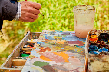Male hand of artist, palette with paints and canvas in nature outdoors and greenery background