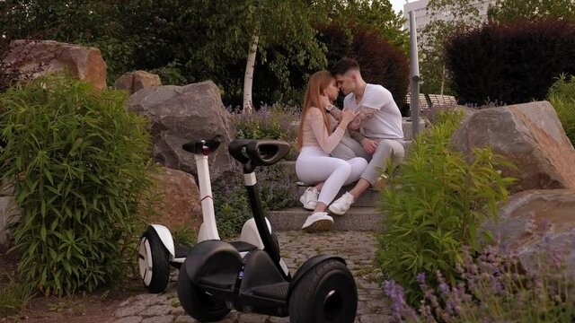 Loving couple are sitting on stone stairs in the park in summer, their gyro scooters are standing nearby. Teenagers riding hoverboards in the park, an active date for a couple in love.