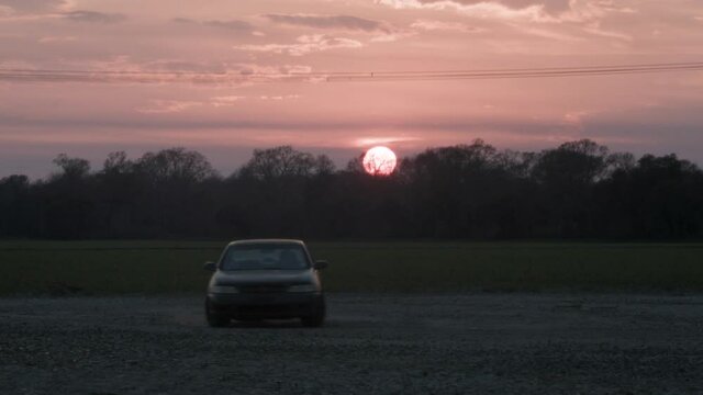 Old Car Drives On Gravel Lot Next To Tractor and Grassy Field During Red Sunset