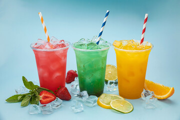 colored lemonades in plastic cups with ice, fruits and berries on a blue background
