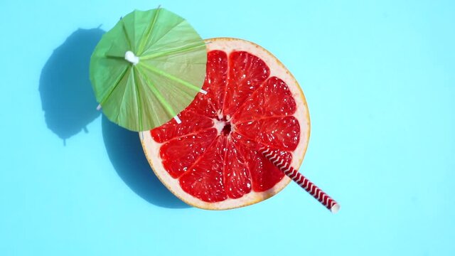 Grapefruit (orange) rotating with a cocktail umbrella in the sunlight on a colored blue background. Fruit juices, relaxation, tropics and relaxation concept.