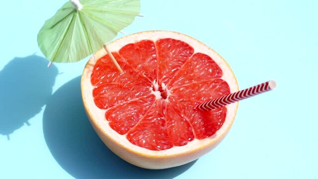 Grapefruit (orange) rotating with a cocktail umbrella in the sunlight on a colored blue background. Fruit juices, relaxation, tropics and relaxation concept.