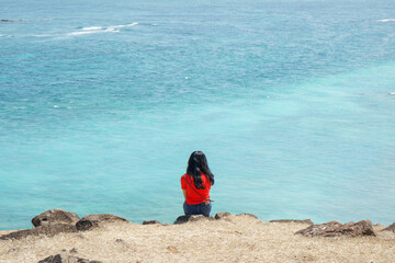 A women in red shirt shit alone on the hill facing the blue water of the sea