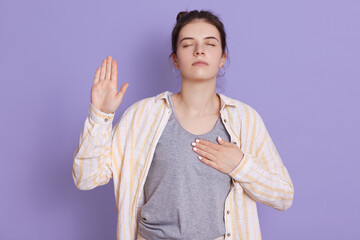 Portrait of hipster woman with dark hair holding hand on heart and raising palm to make oath,...