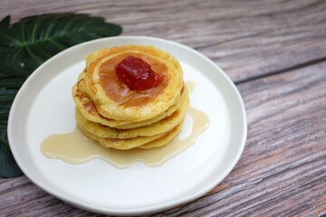 In selective focus a white dish of butter pancake with honey sauce and strawberry jam on top,old wooden table background with blurred green leaves