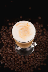 A glass of macciato latte with coffee beans on black mirror background. Cappuccino with milky froth on top with cacao. Selective focus, copy space