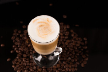 A glass of macciato latte with coffee beans on black mirror background. Cappuccino with milky froth on top with cacao. Selective focus, copy space