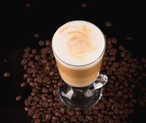 A glass of macciato latte with coffee beans on black mirror background. Cappuccino with milky froth on top with cacao. Selective focus, copy space