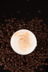 A glass of macciato latte with coffee beans on black mirror background. Cappuccino with milky froth on top with cacao. Selective focus, copy space