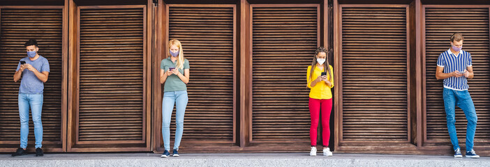 Young people wearing face mask using mobile phone keeping social distance - Millennial friends...