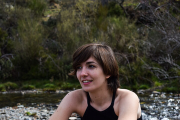 Phot of a young and attractive girl with short hair smiling and standing next to a river with shorts in the nature on a sunny day. Summer clothes