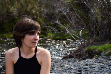 Phot of a young and attractive girl with short hair smiling and standing next to a river with shorts in the nature on a sunny day. Summer clothes