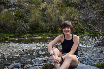 Phot of a young and attractive girl with short hair smiling and standing next to a river with shorts in the nature on a sunny day. Summer clothes