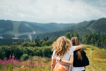 Fototapeta na wymiar Two young girlfriends are standing on a mountain, hugging and enjoying nature with a backpack on flowering slopes on a warm sunny day. Friendship and thirst for entertainment and unity with nature