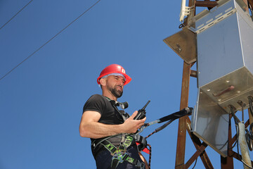 Electrician in red helmet and safety belt with radio station on the electric pole