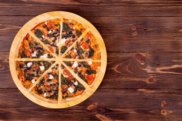 Sliced pizza with cherry tomatoes, spinach, mozzarella, feta, kalamata olive and mushrooms on round wood plate which is on wooden rustic table, top view and space for your text